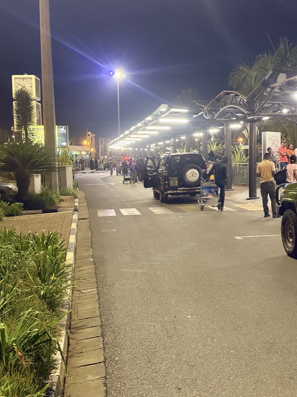 Kigali International Airport arrivals curb at night, with the lit canopy, cars, and luggage carts